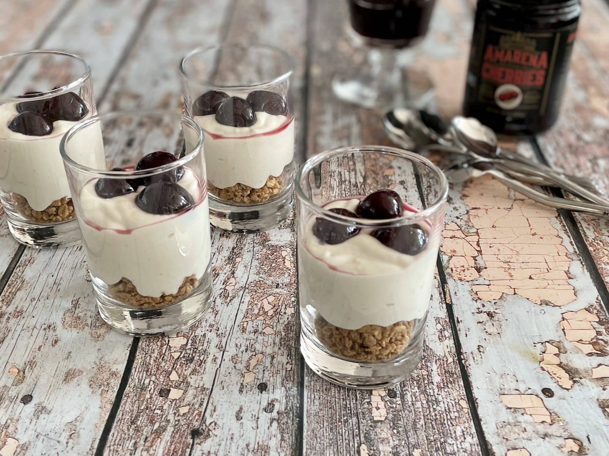 no bake cherry cheesecake in small jars with product amarena-style cherries on wood texture with spoons